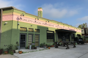 Außenansicht eines grünen Gebäudes mit einer Tafel 'Qualität lokale Versorgung', umgeben von Fenstern, Türen, Lampen, Topfpflanzen, Bäumen, Pfählen, Drähten und einem bewölkten Himmel.