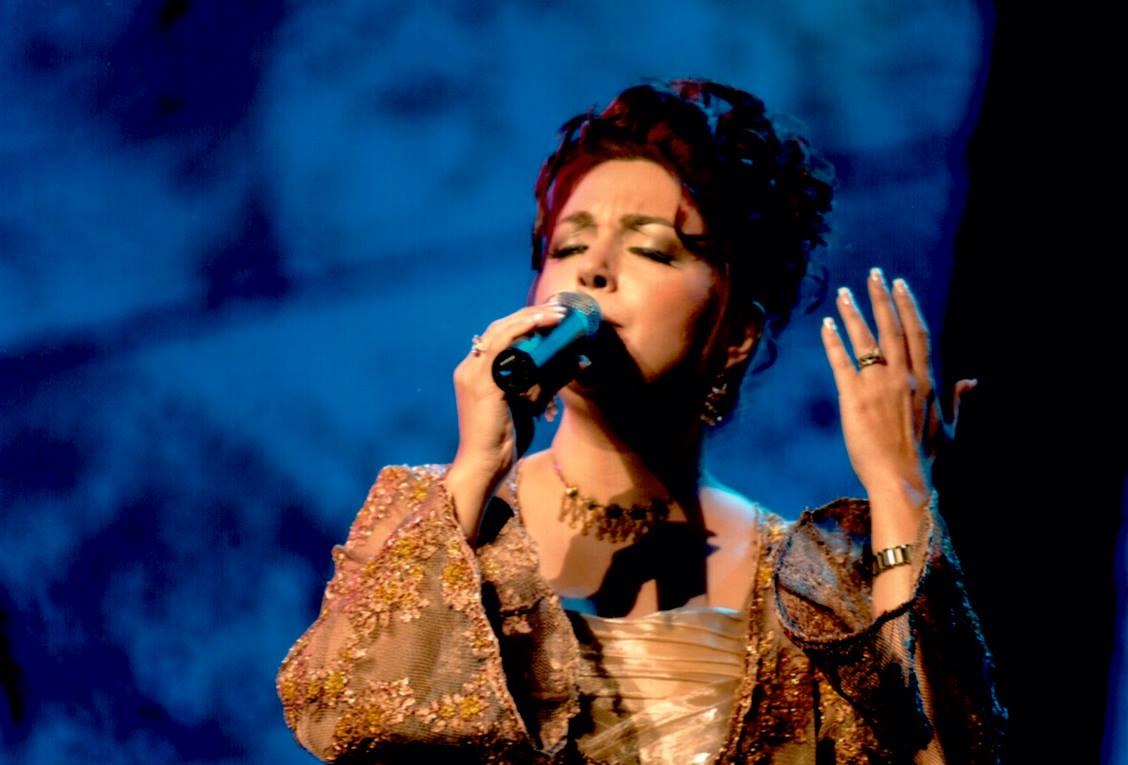Frau in goldenem Kleid, die auf der Bühne in ein Mikrofon singt, mit blauem und schwarzem Hintergrund.