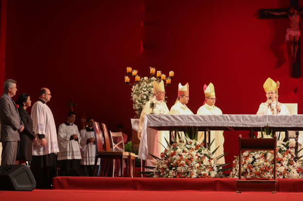 Vier Personen mit Kerzen an Tischen, eine hält ein Mikrofon, Stühle, ein Lautsprecher, Blumen und heilige Kreuze an der Wand im Hintergrund.