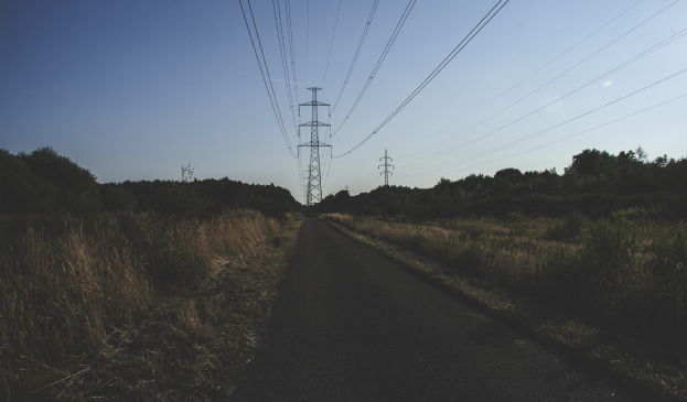 Road with overhead power lines, bordered by green grass and trees, leading to tall transmission towers against a clear blue sky.