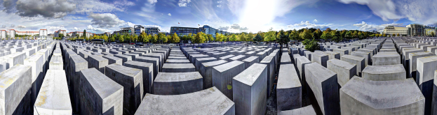 Ein großes Betongebilde namens Holocaust-Mahnmal für die ermordeten Juden Europas in Berlin, Deutschland, umgeben von Bäumen und Gebäuden, mit Fahrzeugen auf der Straße im Vordergrund und einem bewölkten Himmel im Hintergrund.