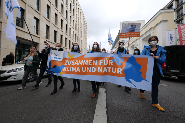 Eine Gruppe von Menschen mit Masken, die eine Tafel mit der Aufschrift "Zusammen für Klima und Natur" tragen, geht eine Straße entlang, mit Fahrzeugen und Gebäuden auf beiden Seiten und Bäumen im Hintergrund unter einem klaren blauen Himmel.