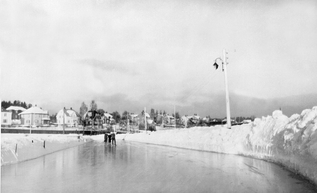 Ein Schwarz-Weiß-Foto von Menschen, die auf einer Eisbahn Schlittschuh laufen, umgeben von Schnee, Pfählen, Lichtern, Drähten, Häusern, Bäumen und Bergen im Hintergrund unter dem Himmel.