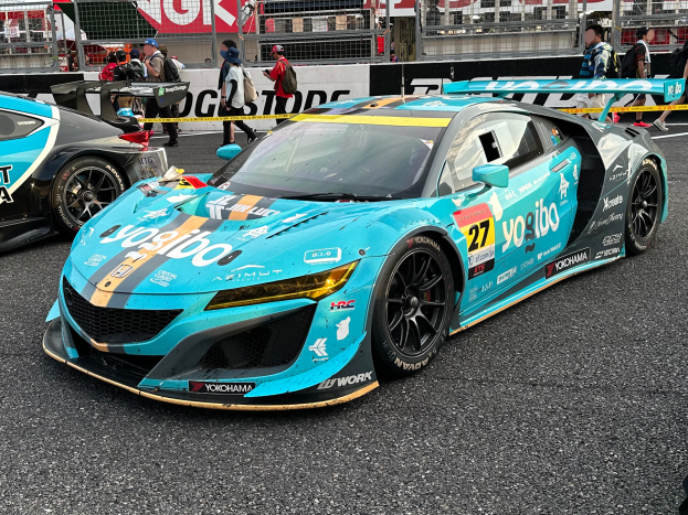 Eine Gruppe von Autos auf einer Rennstrecke geparkt mit Menschen, Geländern, Pfosten und einem Gebäude im Hintergrund und Text unten lesend "Yokohama Acura NSX GT3".