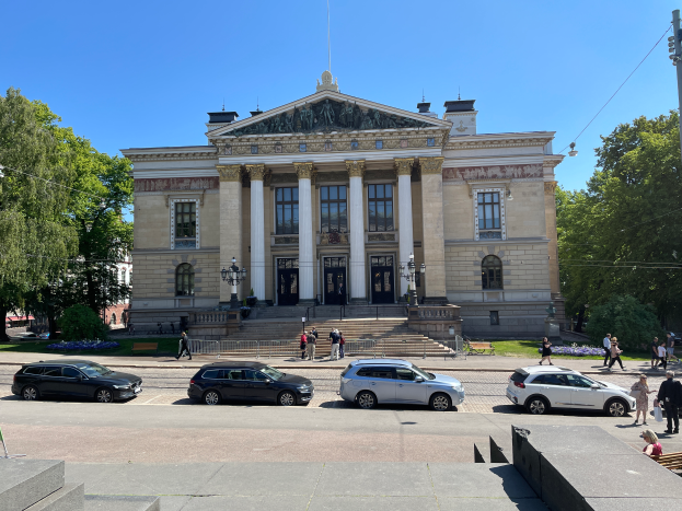 Das schwedische Parlament in Stockholm, Schweden, mit zahlreichen geparkten Autos, Bäumen, Straßenlaternen, Strommasten, Stromkabeln, Kraftfahrzeugen auf der Straße, Fußgängern auf dem Gehweg, Absperrpoller, Büschen, Gras und einem klaren blauen Himmel.