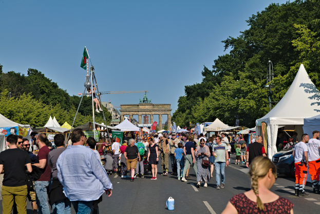 Eine Menschenmenge, die eine Straße mit Zelten, Fahrzeugen und Bäumen entlanggeht, im Hintergrund ein Tor und ein klarer blauer Himmel, sowie links Poles mit Fahnen, die wahrscheinlich das Oktoberfest in München, Deutschland, darstellen.