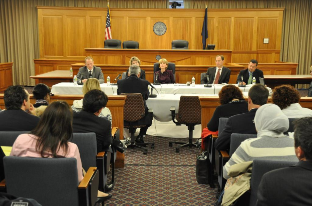 Ein Tagungsraum mit einer Gruppe von Menschen, zwei ausländischen Flaggen, einem orangefarbenen Tisch mit Wasserflaschen und einem Mikrofon.