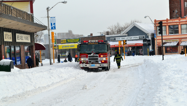 Feuerwehrfahrzeug im Schnee vor Gebäuden geparkt, mit Geschäften und Laternenmasten links, einer Ampel in der Mitte und Schnee auf dem Boden.