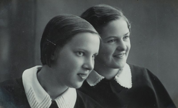 Zwei junge Frauen in Schwarz und Weiß, eine im weißen Kleid und die andere im schwarzen Kleid, lächeln und sehen sich an mit einem leicht unscharfen, nostalgischen Hintergrund.