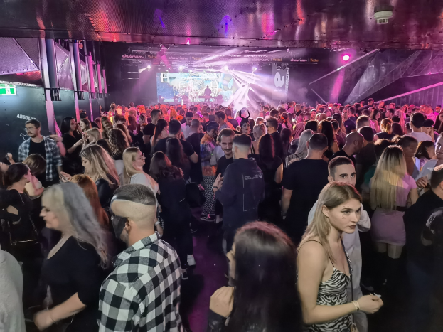 Eine große Gruppe von Menschen in einem Nachtclub mit Deckenbeleuchtung und Säulen, die an einem Absolut Nightlife-Event teilnehmen, mit einem Bildschirm im Hintergrund.