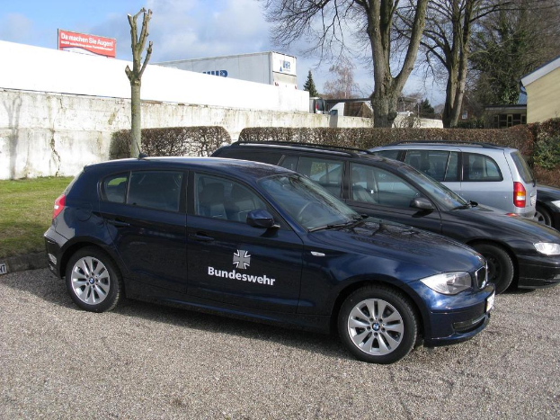 Mehrere Autos sind auf einem von grünem Gras, Büschen und Bäumen umgebenen Weg geparkt. Im Hintergrund sind Gebäude, ein Schild mit Text und ein Logo zu sehen.