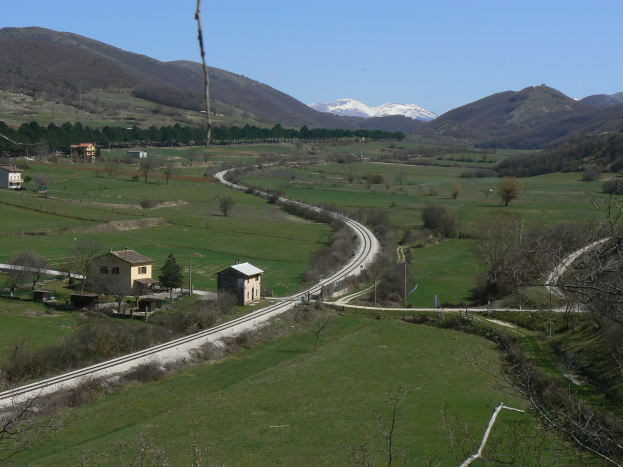 Ein kleines Dorf in einer grünen Talsenke gelegen, umgeben von Bäumen, Pflanzen, Gras und Häusern, mit einer gewundenen Straße, schneebedeckten Bergen im Hintergrund und einem klaren blauen Himmel darüber.