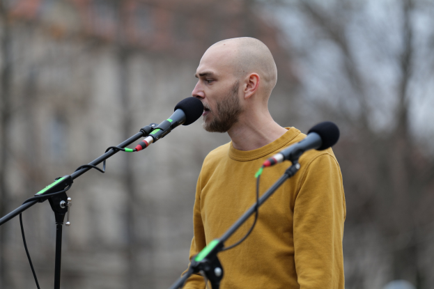 Ein kahlköpfiger Mann in einem gelben T-Shirt singt in ein Mikrofon mit einem unscharfen Hintergrund.