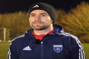 Ein Mann in einer blauen Jacke und Mütze steht auf einem Fußballfeld, umgeben von Bäumen und einem Tor im Hintergrund, während eines Trainings.