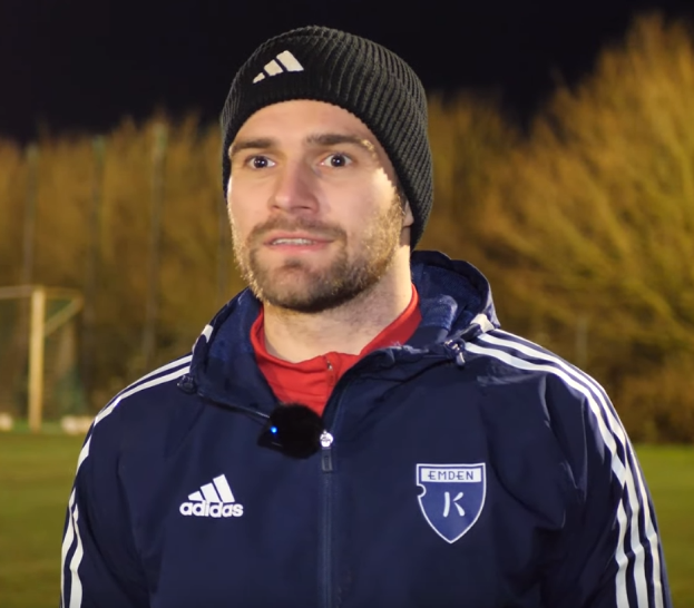 Ein Mann in einer blauen Jacke und Mütze steht auf einem Fußballfeld, umgeben von Bäumen und einem Tor im Hintergrund, während eines Trainings.