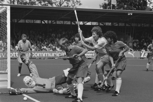 Schwarzes Bild einer Gruppe von Frauen, die Feldhockey spielen, mit Helmen und Stöcken, einem Torpfosten links, Zuschauern in den Rängen, Bannern, einem Schuppen, Bäumen und einer Flagge im Hintergrund.