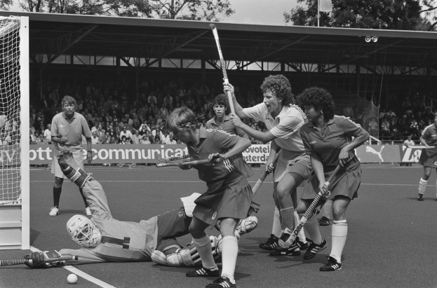 Schwarzes Bild einer Gruppe von Frauen, die Feldhockey spielen, mit Helmen und Stöcken, einem Torpfosten links, Zuschauern in den Rängen, Bannern, einem Schuppen, Bäumen und einer Flagge im Hintergrund.