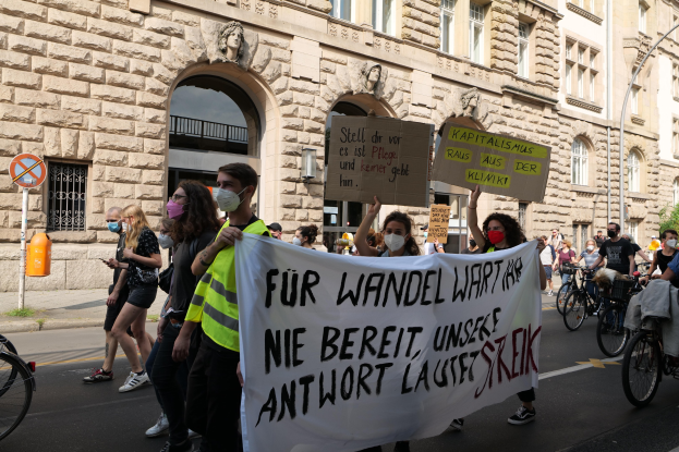 Eine Gruppe von Menschen marschiert bei einer Protestaktion gegen die Anti-Kriegsbewegung in Berlin, einige halten Schilder und andere fahren Fahrräder, im Hintergrund ist ein Gebäude mit Rundbogenfenstern, Säulen und Skulpturen, Bäumen und einem Laternenpfahl zu sehen.