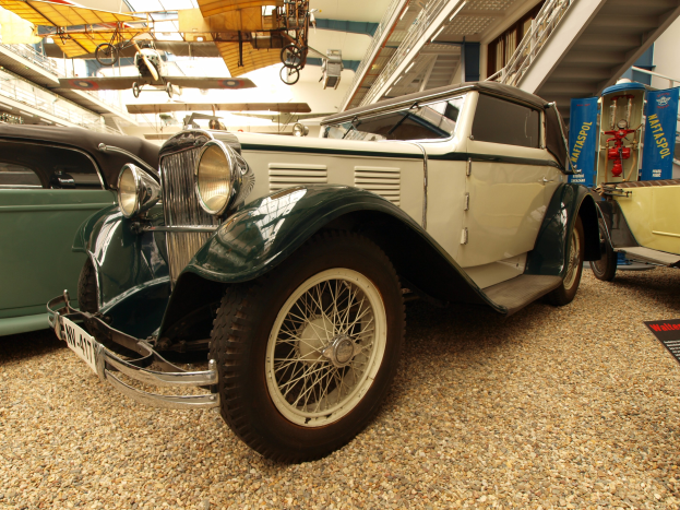 Ein Oldtimer-Auto in einem Museum zwischen anderen Fahrzeugen, mit einer Tafel auf der rechten Seite und Flugzeugen an der Decke im Hintergrund.
