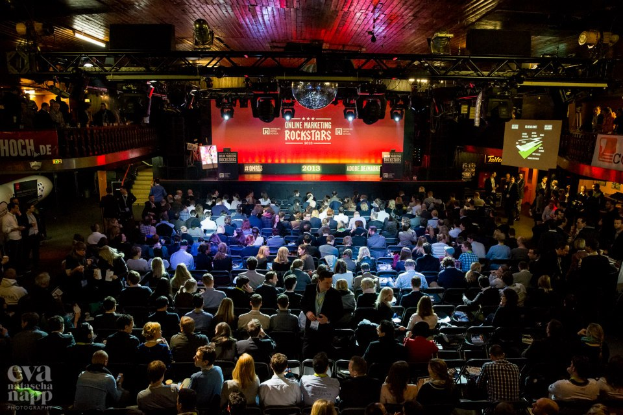 Eine große Menschenmenge sitzt auf Stühlen vor einer Bühne mit Bildschirmen und Bannern, die von Scheinwerfern beleuchtet wird und von Absperrungen umgeben ist, und erwartet gespannt den Beginn eines Online-Marketing-Rockstars-Events, mit einem Wasserzeichen in der linken unteren Ecke.