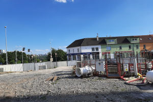 Eine Baustelle mit verstreuten Rohren und Geräten, umgeben von Gebäuden, Laternen, Verkehrszeichen, einem Zaun, Bäumen und einem bewölkten Himmel.