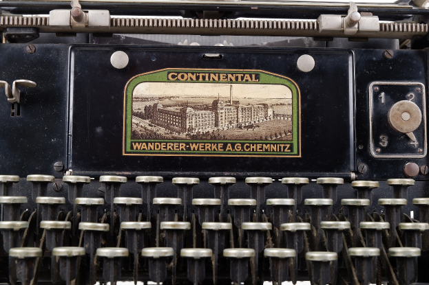 Eine alte schwarze Schreibmaschine mit einem hellgelben Etikett, auf dem "Continental Wanderer Werke A.G. Chemnitz" steht und einer weißen Tastatur.