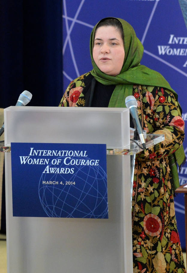 Eine Frau steht an einem Podium mit einem Mikrofon, eine Tafel am Podium befestigt und ein Tisch rechts daneben, mit einem Banner im Hintergrund mit der Aufschrift 'International Women of Courage Awards 2014'.