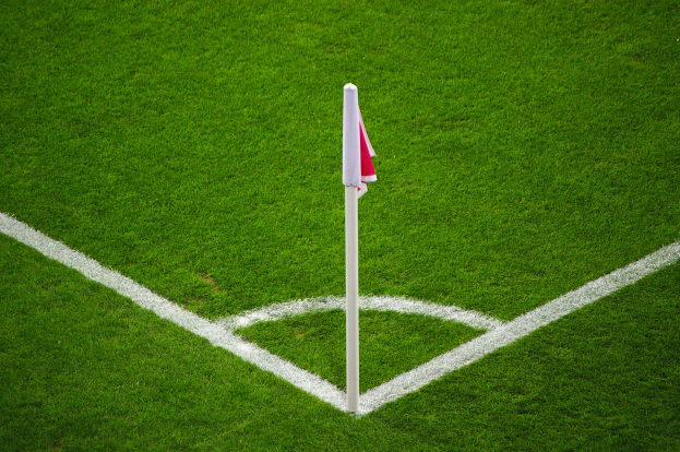 Ein Fussballfeld mit grünem Rasen, weißen Begränzungslinien und einem weißen Pfosten mit einer rot-weißen Flagge auf dem zentralen Strafpunkt.