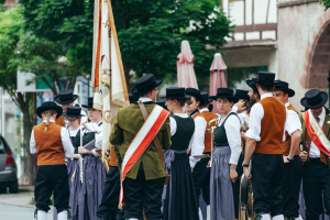 Eine Gruppe von Menschen in traditioneller bayrischer Tracht marschiert auf einer Straße, einige halten Musikinstrumente und Fahnen, im Hintergrund sind Bäume, Gebäude und ein Auto zu sehen.