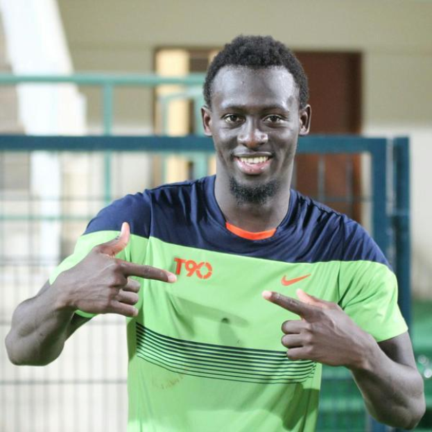 Ein Mann in einem grünen und blauen Hemd, der als Emmanuel Adebayor identifiziert wurde, zeigt mit einem Lächeln auf die Kamera, mit einem unscharfen Geländer, Wand und Tür im Hintergrund.