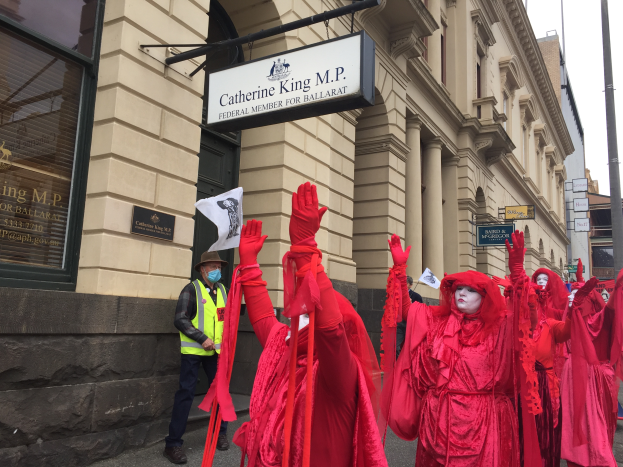 Eine Gruppe von Menschen in roten Kostümen geht eine Straße entlang während eines Ballarat-Faschingsumzugs, mit einer Person in einer grünen Jacke und einem Hut, die ein Papier in der linken Hand hält, mehreren Brettern, die an einem Pfahl vor einem Gebäude befestigt sind, und dem Himmel, der oben im Bild zu sehen ist.