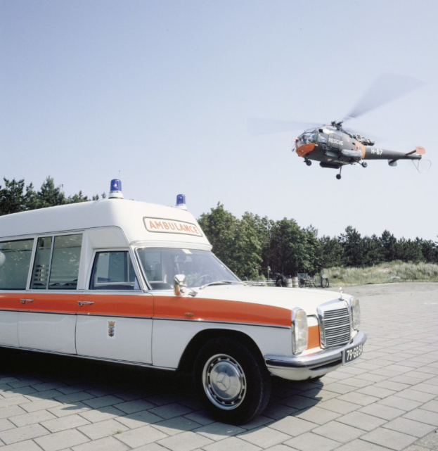 Ein Rettungswagen steht vor einem Hubschrauber, umgeben von Bäumen und einem klaren blauen Himmel.
