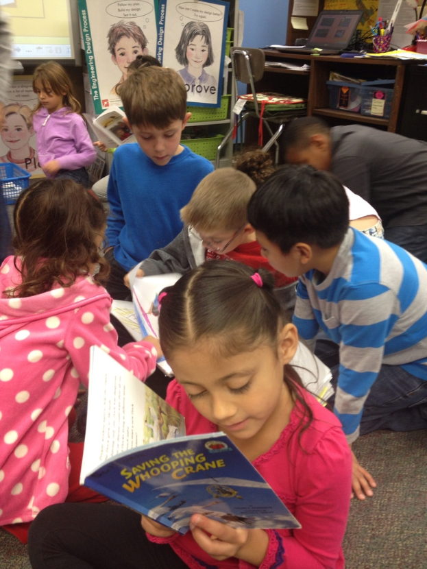 Eine Gruppe von Kindern sitzt auf dem Boden, einige halten B"ucher, mit K"rben, Gegenst"anden, Papieren mit Bildern, einem Laptop und anderen Gegenst"anden auf Regalen und einem Tisch im Hintergrund.