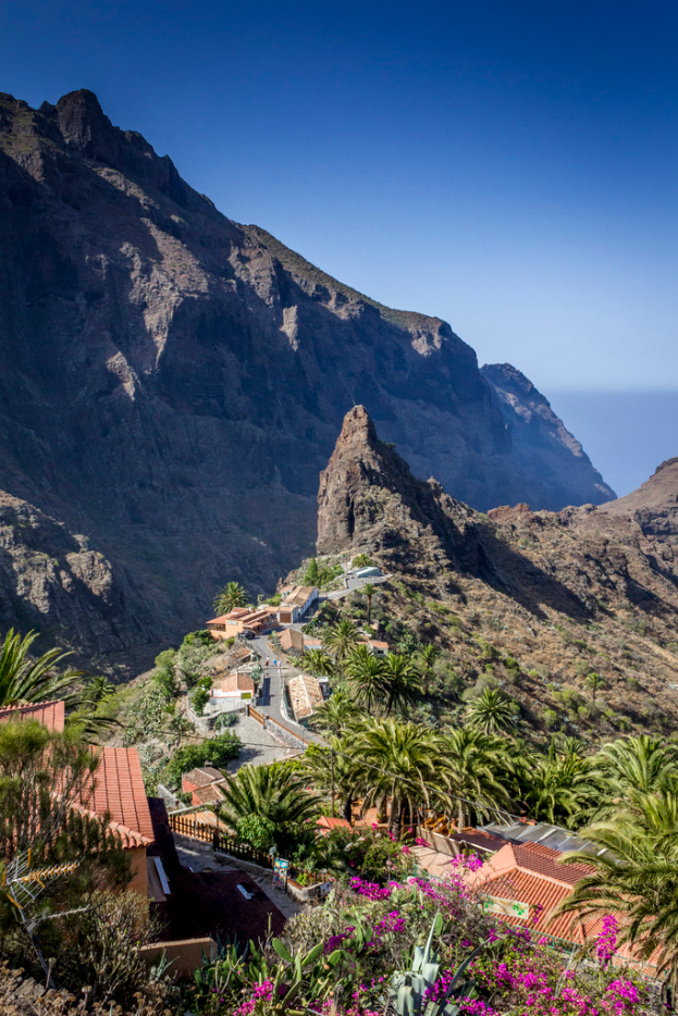 Eine ländliche Szene mit Häusern, Bäumen und Felsen im Vordergrund, einem Hügel im Hintergrund und einem klaren Himmel darüber.