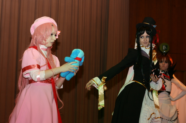 Drei Frauen führen ein Drama auf, wobei eine in einem rosafarbenen Kleid ein blaues Stofftier hält, eine andere in einem schwarzen Kleid nach links schaut und ein brauner Vorhang im Hintergrund zu sehen ist.