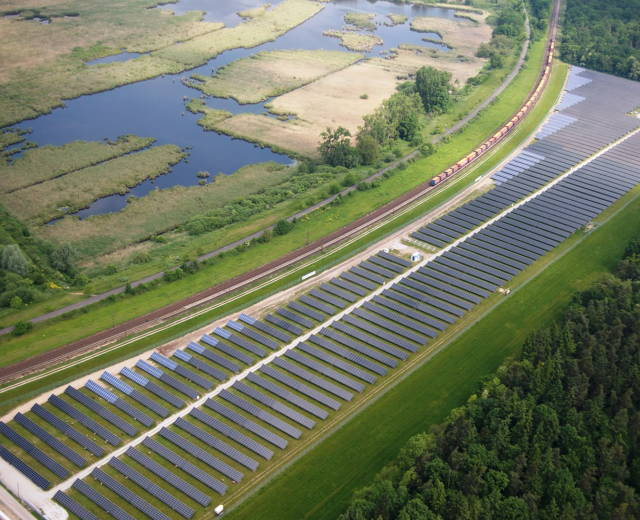 Luftaufnahme eines Solarparks mit Panelen in einem Feld, umgeben von Bäumen, Gras, Pflanzen und Wasser, mit einem Zug auf einer näheren Eisenbahnschiene.