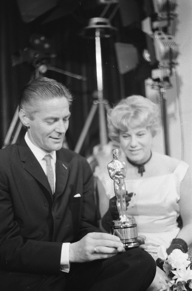 Ein Mann in einem schwarzen Blazer, weißem Hemd und Krawatte hält eine Oscar-Statue, während er neben einer Frau in Abendgarderobe sitzt, die einen Blumenstrauß hält, vor schwarzem Hintergrund in einem Schwarz-Weiß-Bild.