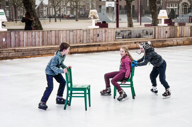 Kinder beim Skifahren im Vordergrund eines Spielplatzes mit drei Kindern und zwei Stühlen in der Mitte sowie Gebäuden, Bäumen, Bänken, Pfosten und einem Basketballfeld im Hintergrund.