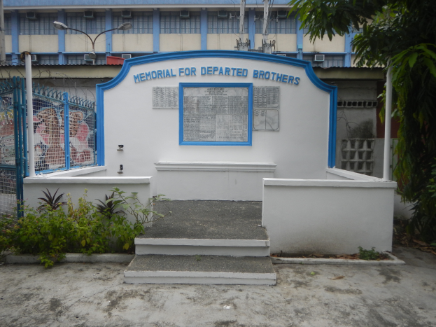 Ein Denkmal f├╝r verstorbene Br├╝der steht vor einem Geb├Ąude mit einem blauen und wei├čen Schild, umgeben von einem Metallzaun, Pflanzen, einer Treppe, Masten, Dr├Ąhten und einem Baum, unter einem bew├Âlktem Himmel.