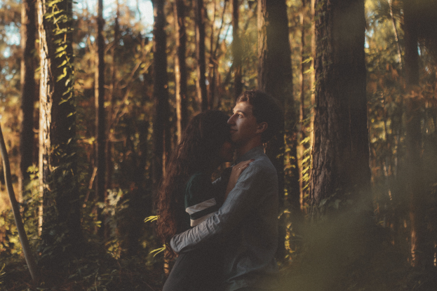 Ein Mann und eine Frau umarmen und küssen sich in einem bewaldeten Gebiet mit hohen Bäumen und üppiger Vegetation, unter einem sichtbaren Himmel, während ihrer Verlobungsfotoshoot.