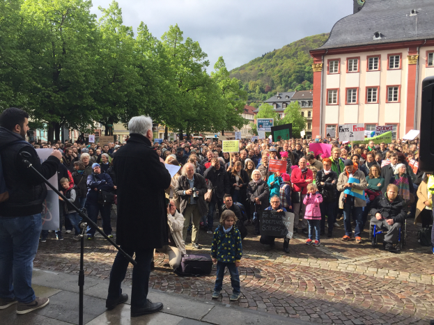 Ein Mann steht vor einer Menge mit Schildern, spricht in ein Mikrofon in Heidelberg, Deutschland, mit Bäumen, Gebäuden, einem Hügel und einem bewölktem Himmel im Hintergrund.