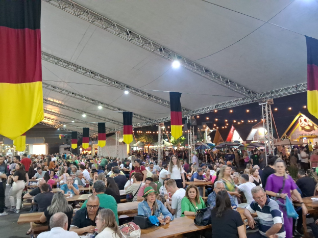 Große Gruppe von Menschen an langen Tischen unter einem Zelt auf dem Oktoberfest in München, Deutschland, mit Taschen und Gegenständen auf den Tischen und festlicher Dekoration im Hintergrund.