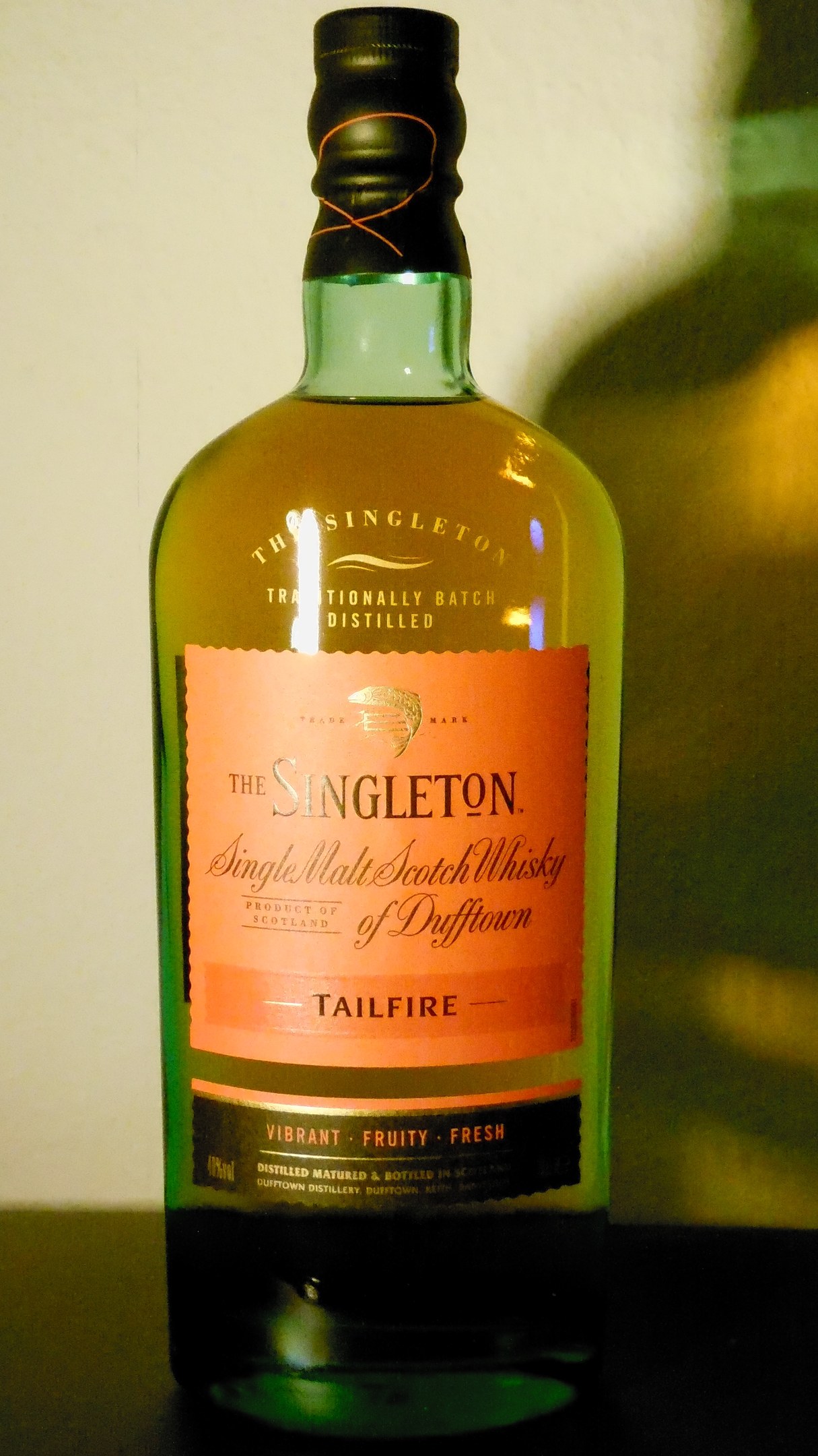 Eine tiefe bernsteinfarbene Flasche Singleton of Dufftown Tailfire Single Malt Scotch Whisky auf einem Tisch, mit einem schwarzen Etikett, das ein Wappen mit einem Löwen und einem Schloss zeigt.