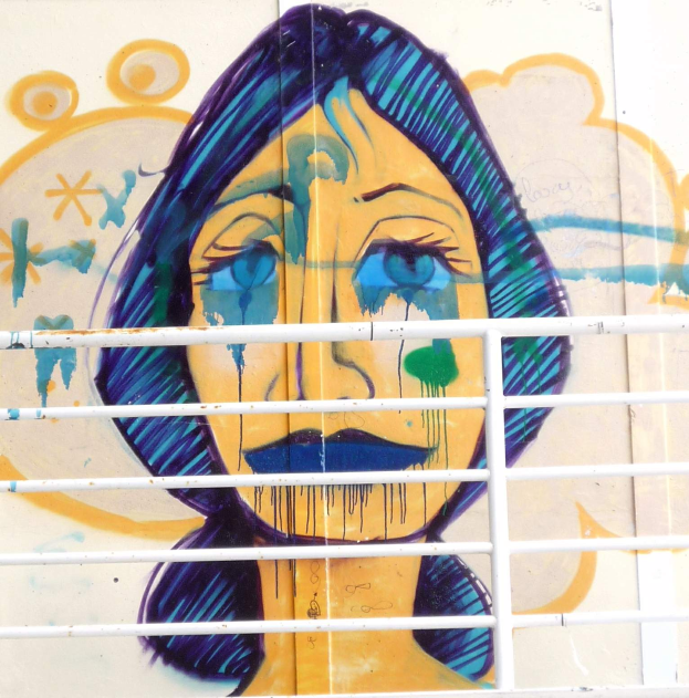 Ein Wandbild eines traurigen Frauengesichts mit geschlossenen Augen und geöffneten Lippen, das auf einem Gebäude in Berlin, Deutschland, gemalt ist, mit einem Gitter im Vordergrund.