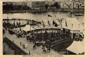 Ein altes Schwarz-Weiß-Foto einer Menge, die sich um einen Karneval mit Zelten, Fahnen, Mäständen und Pferden im Vordergrund und Gebäuden, Bäumen und Wasser im Hintergrund versammelt hat, mit Text am unteren Bildrand.