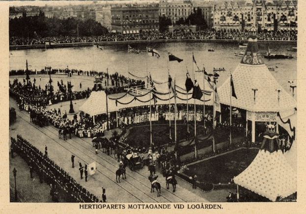 Ein altes Schwarz-Weiß-Foto einer Menge, die sich um einen Karneval mit Zelten, Fahnen, Mäständen und Pferden im Vordergrund und Gebäuden, Bäumen und Wasser im Hintergrund versammelt hat, mit Text am unteren Bildrand.