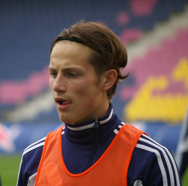 Ein junger Mann in einer orangefarbenen Weste steht auf einem Fußballfeld und streckt die Arme aus, mit entschlossener Miene.