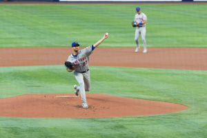 Ein Baseballspieler in einem Sportoutfit, einschließlich einer Mütze und Handschuhen, wirft einen Ball auf einem Feld mit einer Texttafel im Hintergrund.