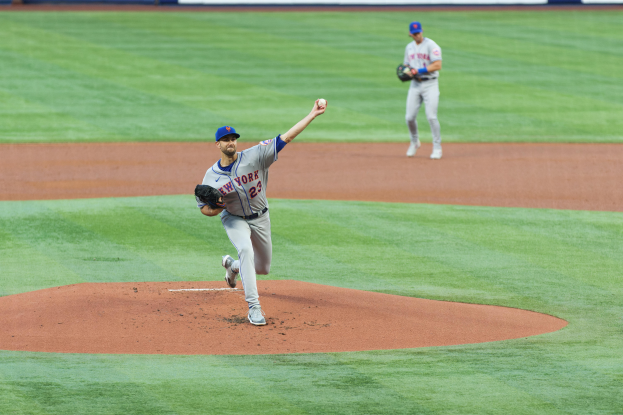 Ein Baseballspieler in einem Sportoutfit, einschließlich einer Mütze und Handschuhen, wirft einen Ball auf einem Feld mit einer Texttafel im Hintergrund.