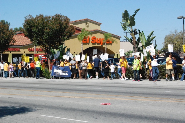 Eine Gruppe von Menschen mit Schildern und B├╝nsten, mit B├╝schen, einem Banner, einem Geb├Ąude mit Namensschildern, Autos und aktuellen Umfragen im Hintergrund.